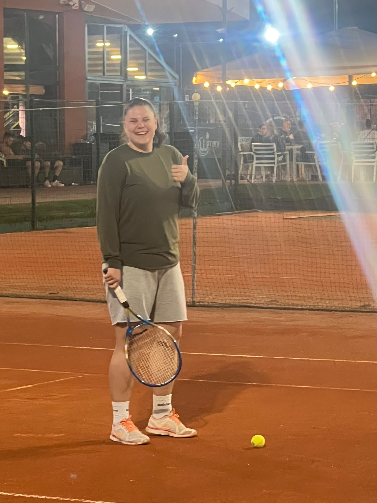 Roxi făcând semn cu degetul mare, cu racheta de tenis pe teren